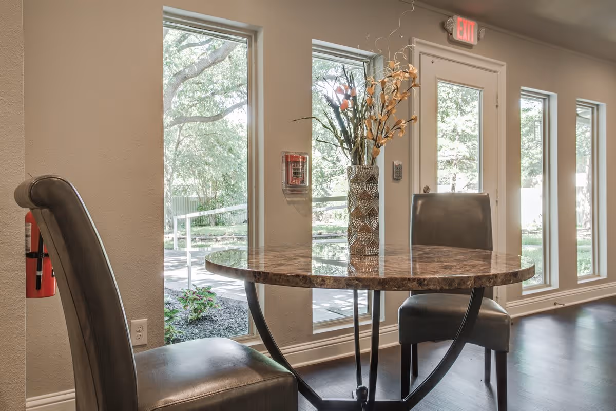 A small round table with a marble-like surface and two gray upholstered chairs in a room with large windows and a glass door showing a green outdoor area. A decorative vase with dried flowers is placed on the table. A fire extinguisher is mounted on the wall to the left, and an exit sign is above the door.