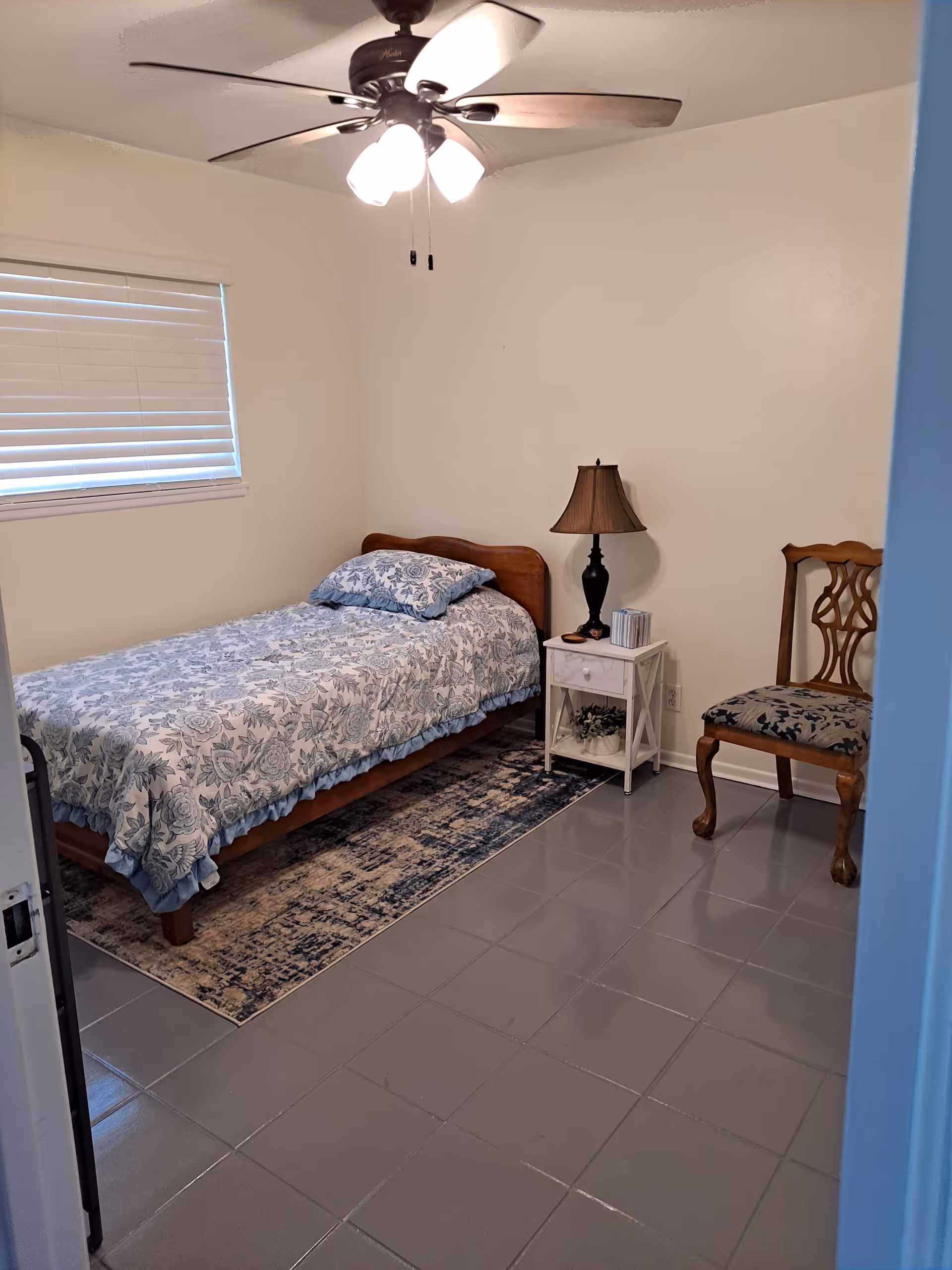 A small bedroom with a single wooden bed covered with a blue and white floral bedspread and matching pillow. Next to the bed is a white nightstand with a black lamp, a box of tissues, and a small plant underneath. A wooden chair with a patterned cushion is placed against the wall. The floor is tiled in gray, and there is a ceiling fan with lights above. A window with closed blinds is on the left wall.