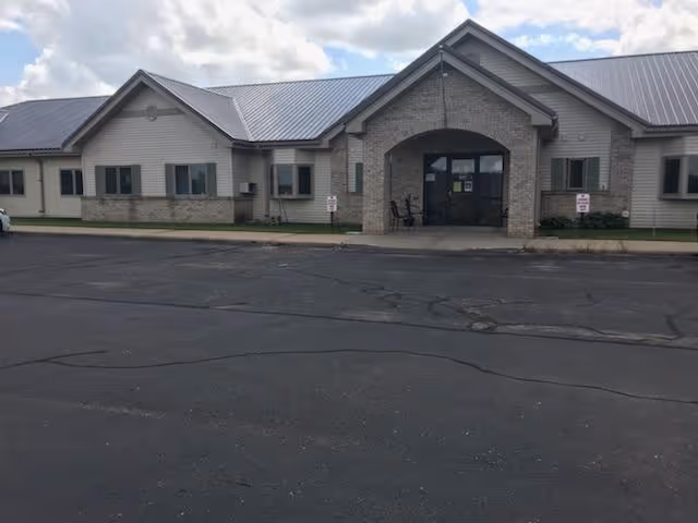 Single-story assisted living building with light brick and siding, a covered main entrance, and a large asphalt parking area under a partly cloudy sky.