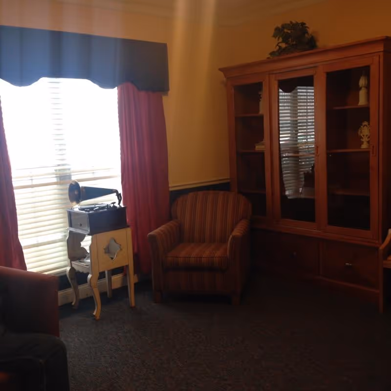A cozy living room area with a striped armchair, a wooden cabinet with glass doors displaying decorative items, a small table holding a vintage gramophone, and a window with closed blinds and red curtains.
