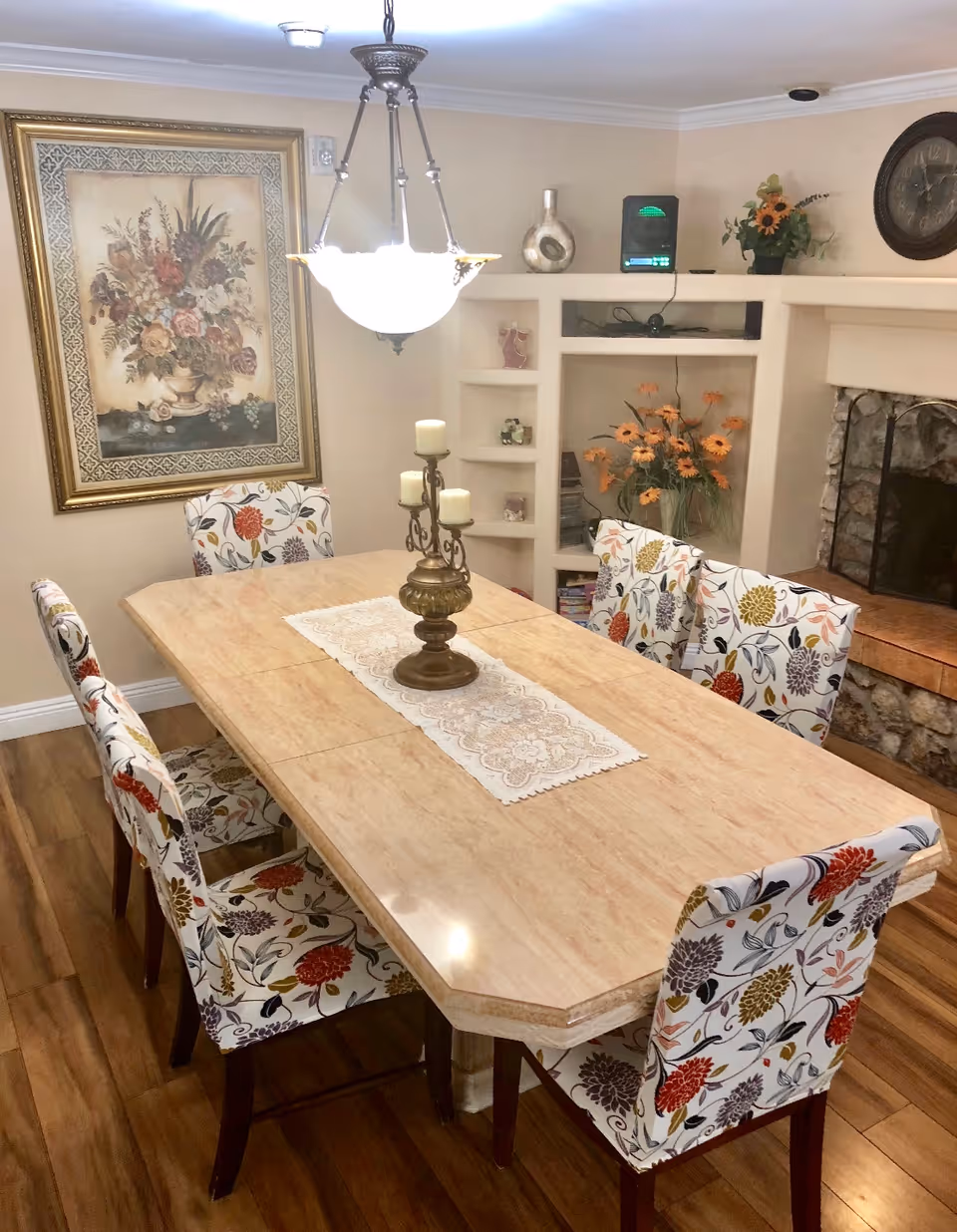 A dining room with a rectangular marble table surrounded by six floral-patterned chairs. A decorative candle holder with three candles sits on a lace runner in the center of the table. The room features wooden flooring, a hanging light fixture above the table, a large framed floral painting on the wall, built-in shelves with decorative items, a vase of orange flowers, and a stone fireplace with a clock above it.