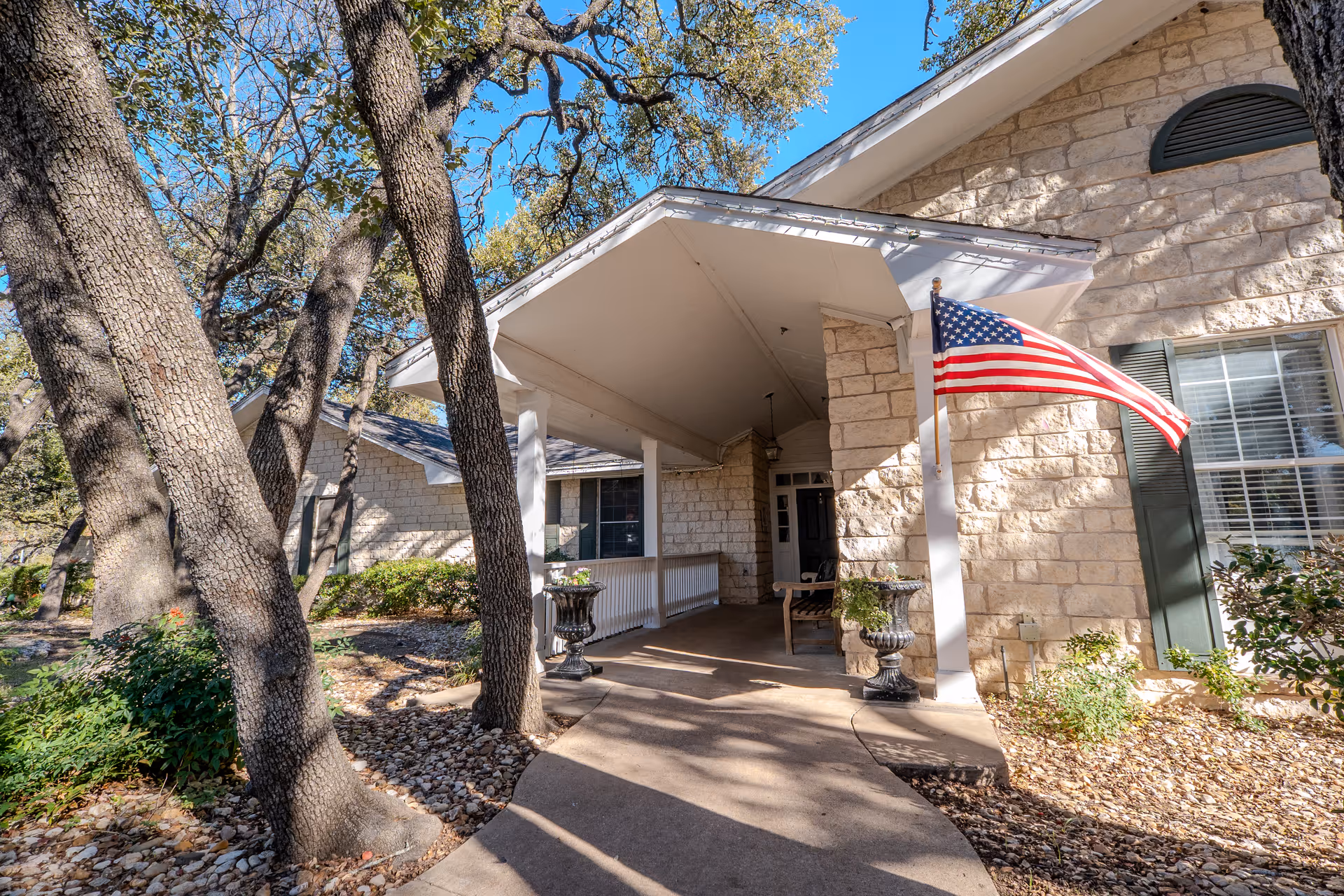 Covered stone building entrance and porch with an American flag and oak trees in front.