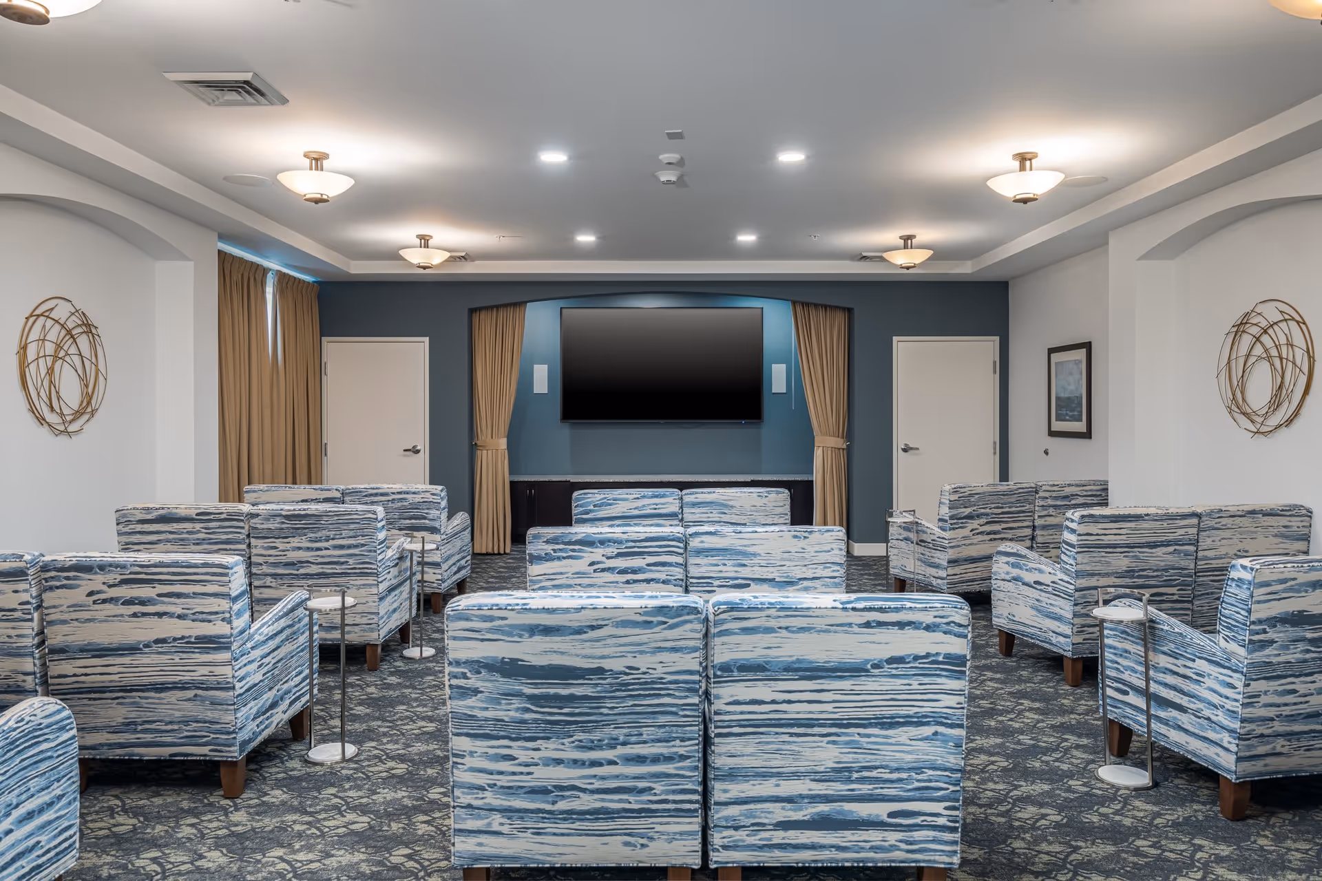 Seating area with patterned armchairs arranged facing a wall-mounted TV in a senior living community common room.