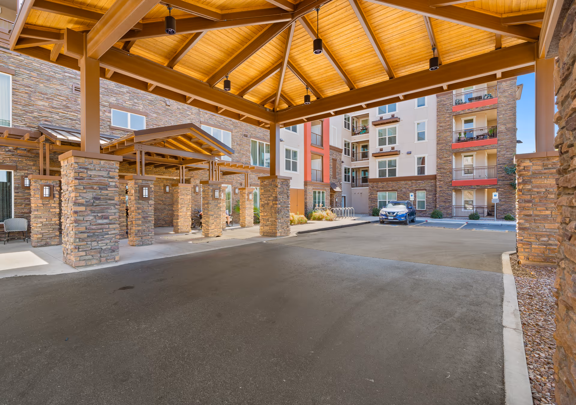 Covered entrance area with stone pillars and wooden ceiling at a senior living facility, with a parking lot and multi-story building in the background.