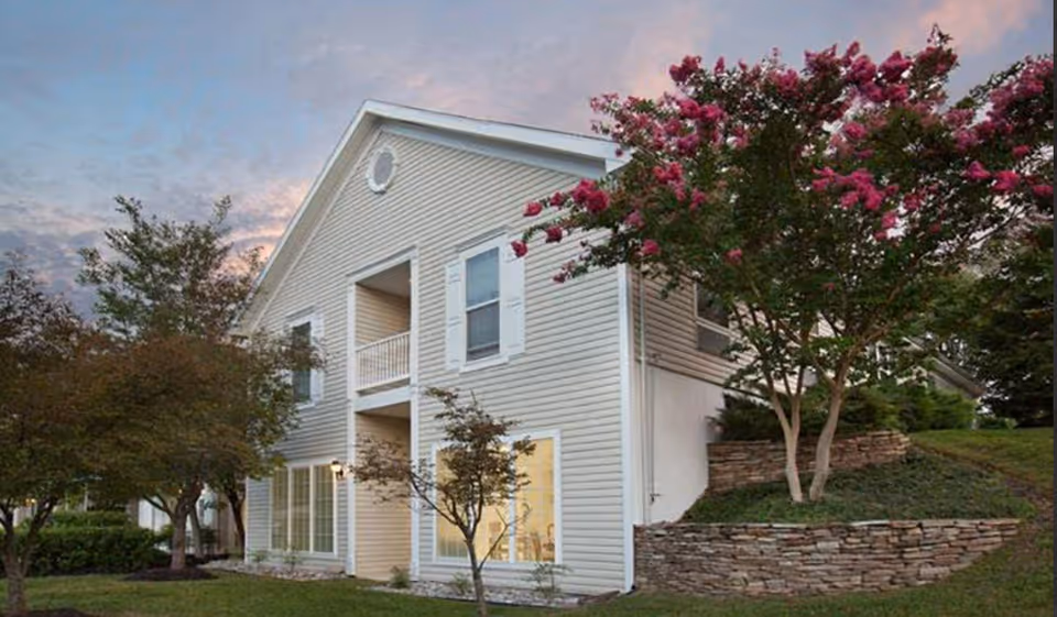 Exterior view of a two-story beige building with white trim at sunset, surrounded by green grass, trees, and a stone retaining wall with blooming pink flowers.
