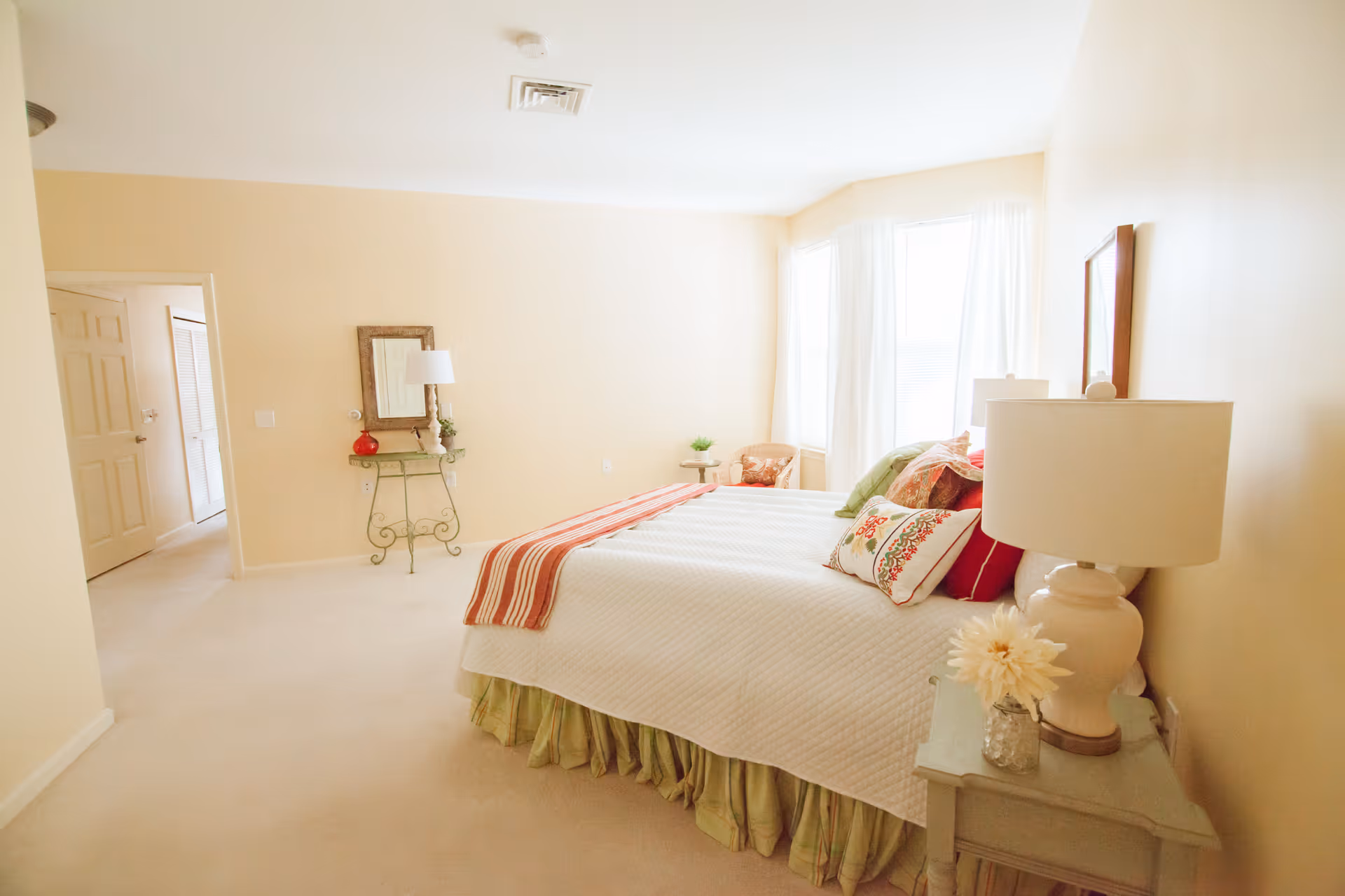 Bright, softly decorated bedroom with a bed dressed in white linens and colorful pillows, a bedside table with a lamp, and a small console table near the doorway.