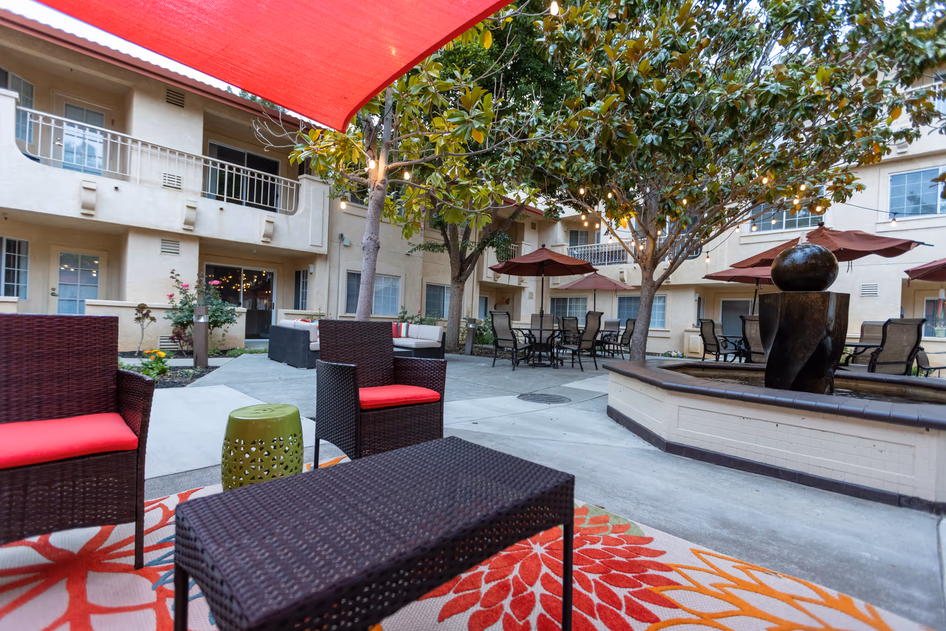 Outdoor courtyard area at Cogir of North Bay Senior Living featuring wicker chairs with red cushions, a wicker coffee table, a colorful floral outdoor rug, several patio tables with umbrellas, trees with string lights, and a water fountain surrounded by a tiled base. The courtyard is enclosed by a multi-story building with balconies and windows.