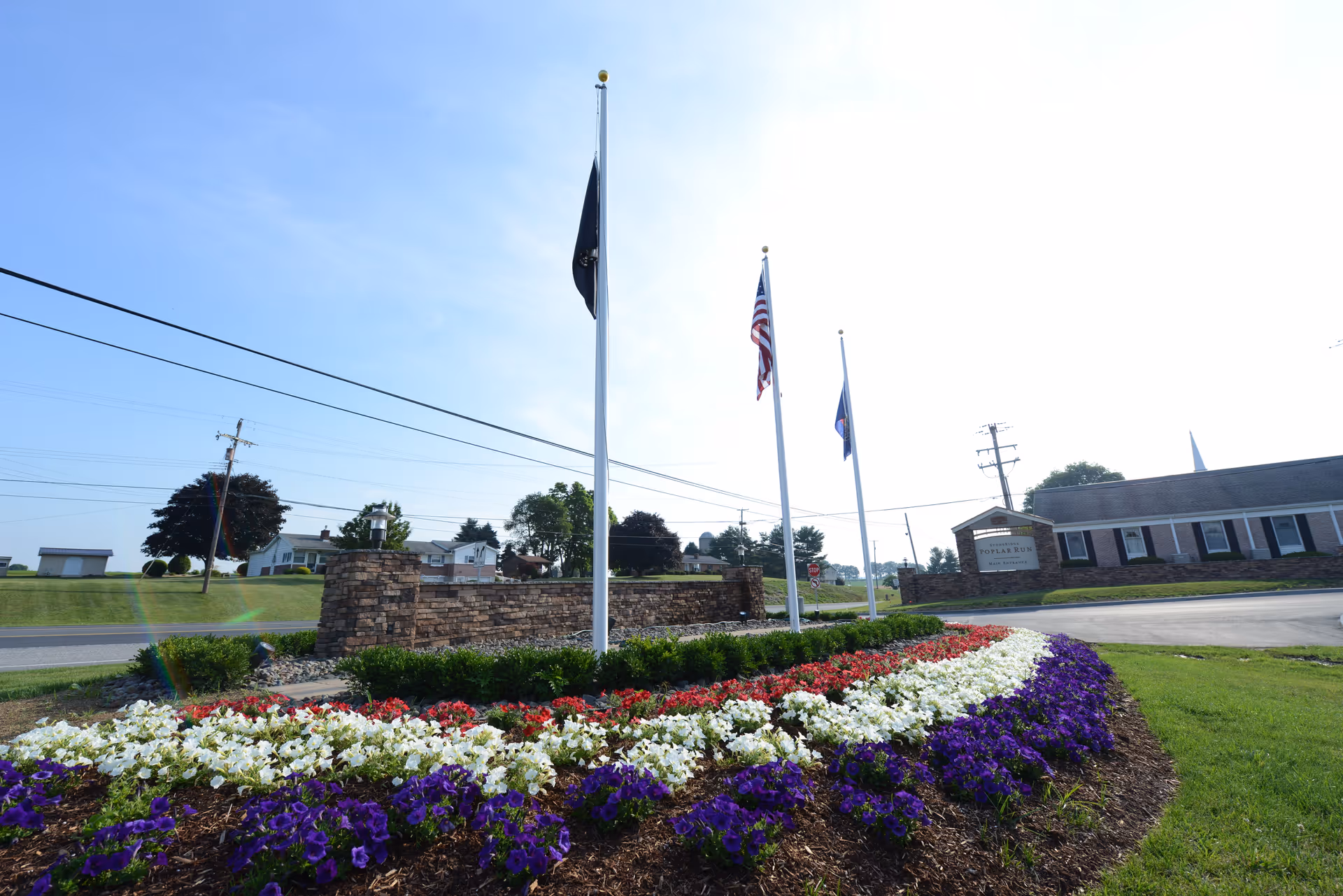 A landscaped entrance area with colorful flower beds in purple, white, and red, three flagpoles with flags, and a stone wall with a sign for Poplar Run a StoneRidge Retirement Living Community. There is a building and a road in the background under a clear blue sky.
