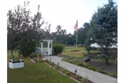 Concrete walkway through grassy landscaped grounds with a small white gazebo, an American flag on a flagpole, trees, and planters.