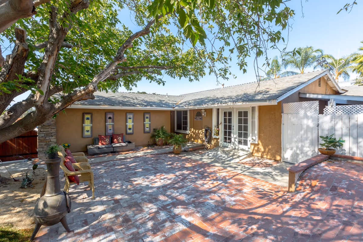 Outdoor patio area at Assisted Living at Golden Way Care of Chino Hills featuring a large tree providing shade, brick flooring, wicker chairs with red cushions, a small sofa with decorative pillows, potted plants, and a beige building with French doors and a welcome sign.