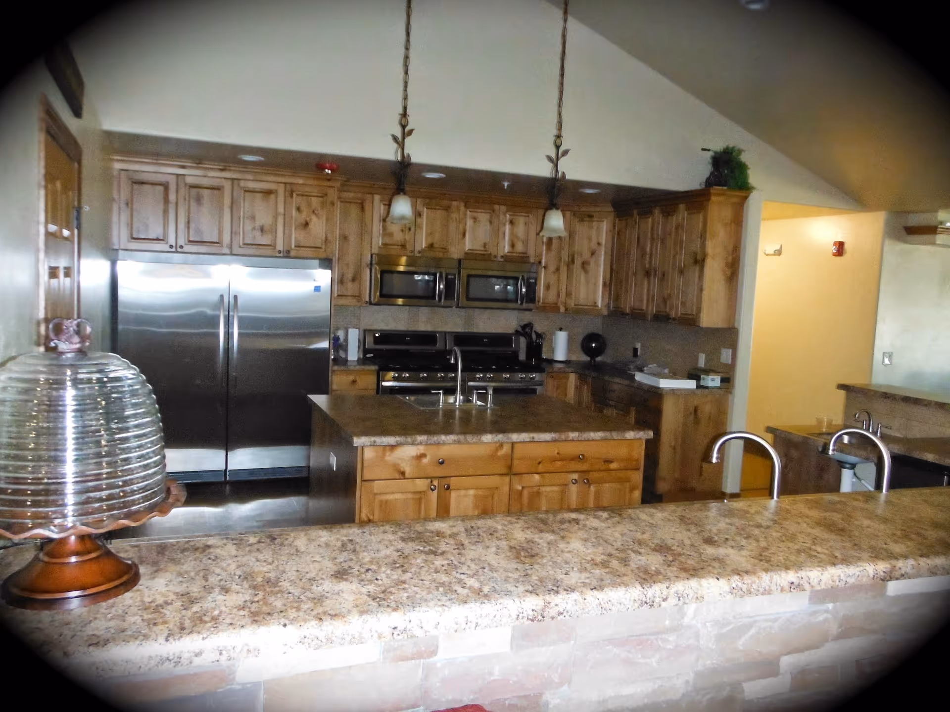 A spacious kitchen with wooden cabinets, a large island with a sink, stainless steel double-door refrigerator, double ovens, and two pendant lights hanging from the ceiling. The countertop is made of a speckled stone material, and there is a glass cake stand on the counter in the foreground.