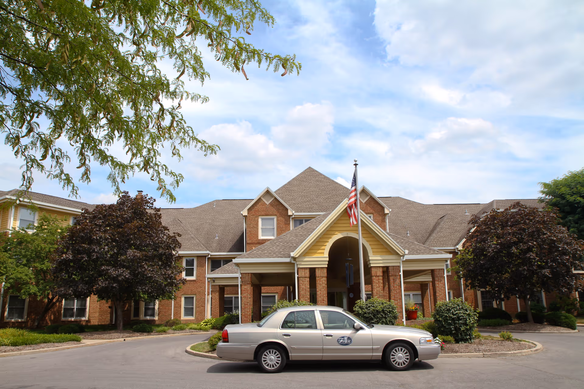 Front entrance of a brick senior living facility with a covered porte-cochere, American flag, landscaping, and a parked sedan.