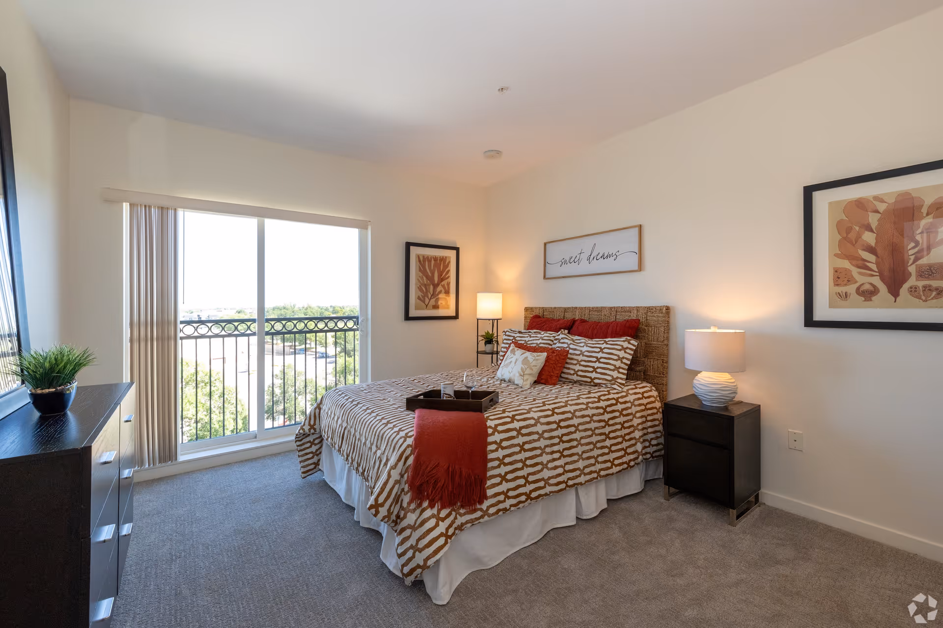 A bright bedroom with a large window and sliding glass door leading to a balcony. The room features a bed with patterned bedding, red and white pillows, a red throw blanket, two framed artworks on the walls, a nightstand with a lamp, a dresser with a small plant, and carpeted flooring.