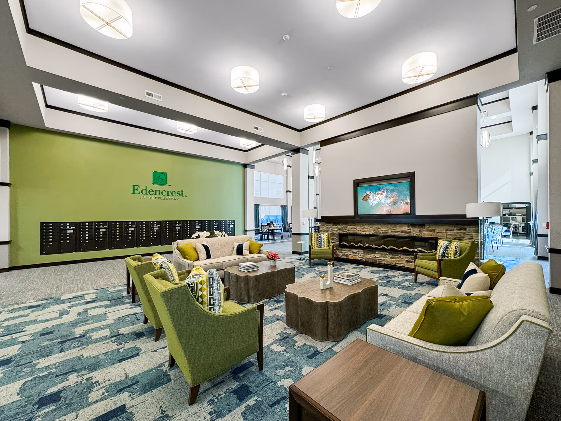 A spacious and modern senior living facility lounge area at Edencrest at Kettlestone featuring comfortable seating with green and beige armchairs and sofas arranged around two uniquely shaped coffee tables. The room has a patterned blue and beige carpet, a stone fireplace with a mounted flat-screen TV above it, and a green accent wall with the Edencrest at Kettlestone logo. The area is well-lit with ceiling lights and has large windows allowing natural light to enter.