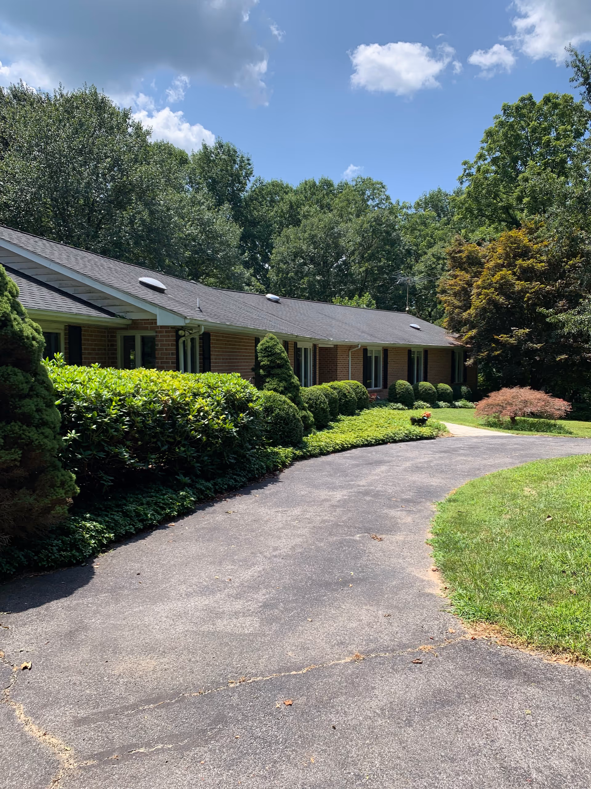 Driveway leading to the front of a single-story brick building surrounded by trimmed shrubs and trees under a partly cloudy sky.