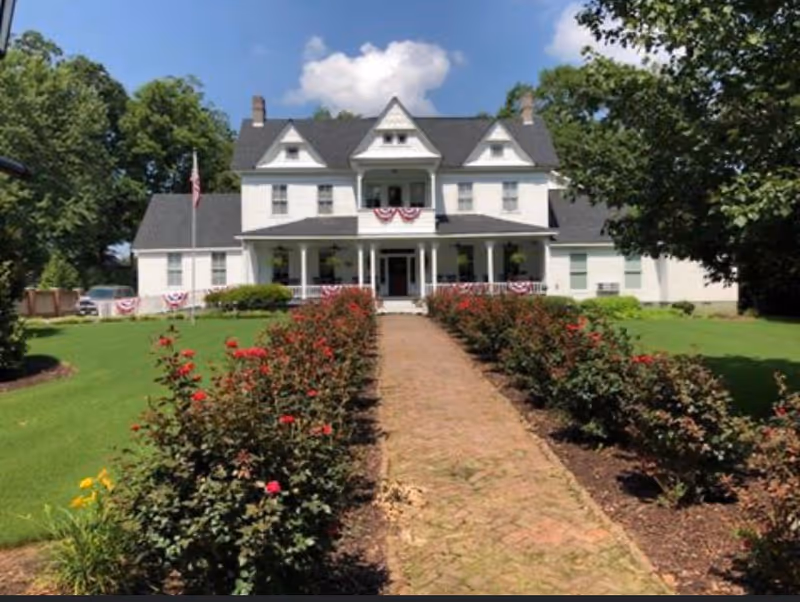 A large white multi-story building with a gray roof, featuring a front porch decorated with red, white, and blue bunting. A brick pathway lined with rose bushes leads up to the entrance. The building is surrounded by green grass and trees under a blue sky with some clouds.