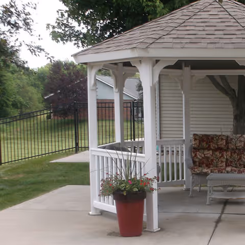 A white wooden gazebo with a shingled roof on a concrete patio. There is a red planter with flowers in front of the gazebo. Inside the gazebo, there is a cushioned bench with floral patterned cushions and a small table. In the background, there is a black metal fence, green grass, trees, and a building with white siding and a brick base.