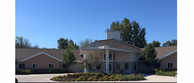 Front exterior of Milan Estates senior living building with a covered entrance, circular driveway, and landscaped bed under a clear blue sky.