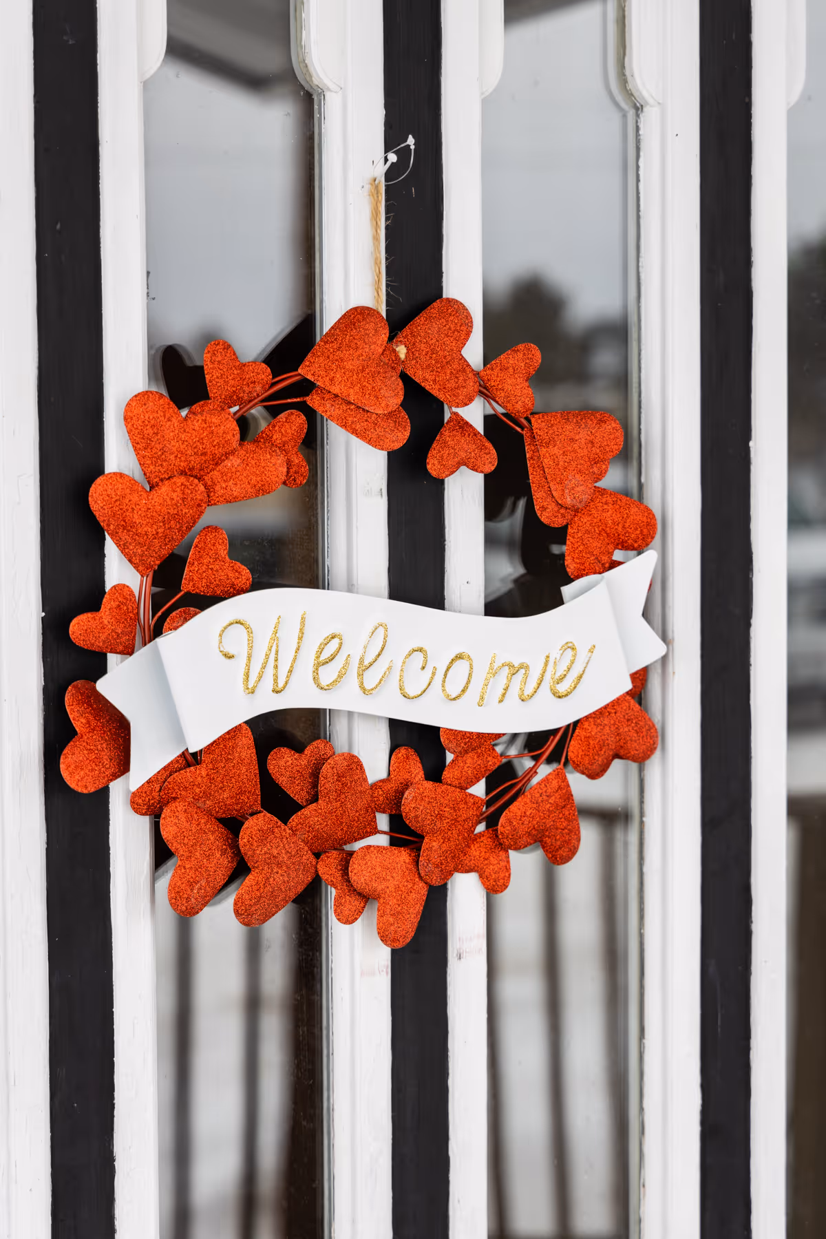A decorative wreath made of red glittery hearts with a white ribbon across the middle that says 'Welcome' in gold glitter, hanging on a black and white door.