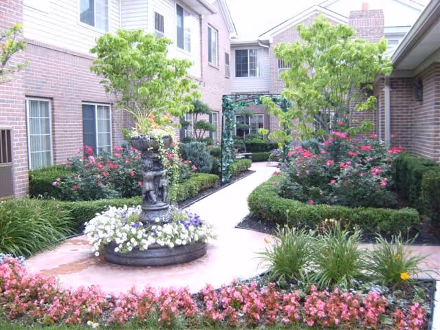 A landscaped courtyard garden at Villa Bella featuring a central stone fountain surrounded by white and purple flowers. The garden is bordered by neatly trimmed bushes, pink flowering plants, and small trees. The courtyard is enclosed by brick buildings with windows, and a paved walkway leads through the garden.