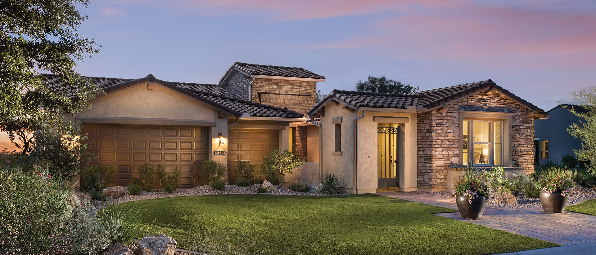 Exterior view of a single-story house at sunset with a well-maintained lawn, stone and stucco facade, two-car garage, and a front entrance with a glass door. The sky is clear with a pinkish hue.