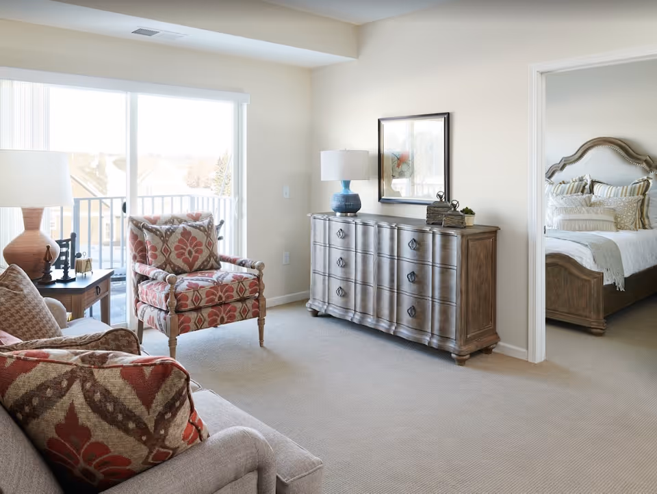 A bright living room area with a patterned armchair and sofa, a wooden side table with a lamp, and a wooden dresser with a blue lamp and decorative items. A doorway leads to a bedroom with a bed featuring a wooden headboard and multiple pillows. Large sliding glass doors open to a balcony with a view outside.