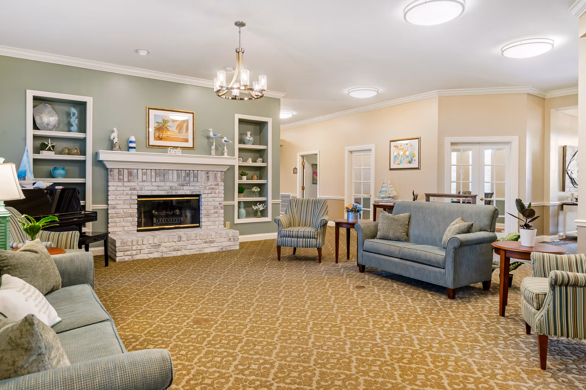 A cozy living room area in a senior living facility featuring a brick fireplace with decorative items on the mantel and built-in shelves on either side. The room has multiple upholstered chairs and sofas in soft green and striped patterns, wooden side tables with plants and flowers, a piano in the corner, and framed artwork on the walls. The carpet has a patterned design, and the ceiling has modern light fixtures.