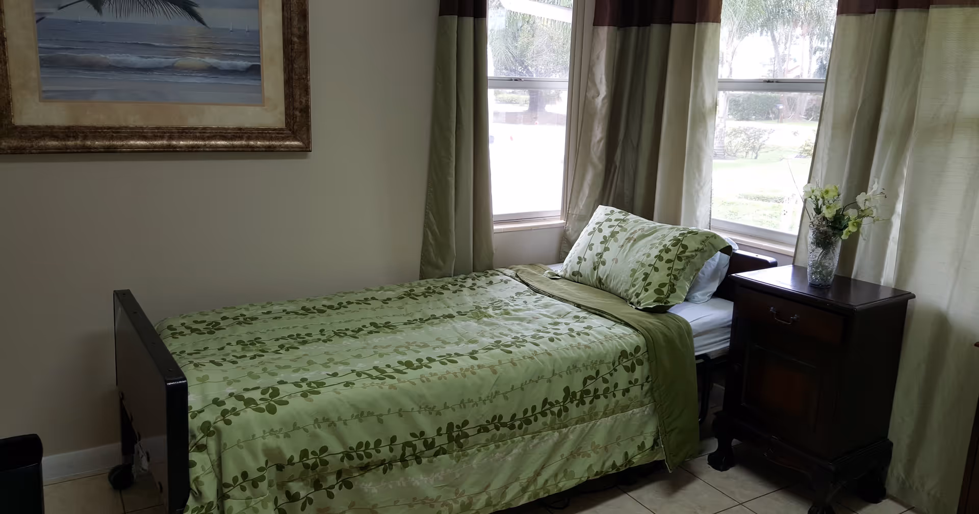 Single bed with green leaf-patterned bedding beside a wooden nightstand and windows with curtains.