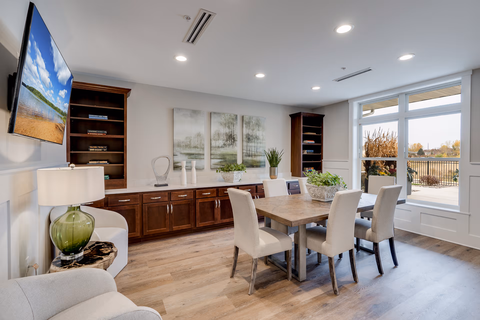 A bright and modern common area with a wooden dining table surrounded by six upholstered chairs. The room features large windows letting in natural light, wooden cabinets with decorative items, two tall bookshelves, a green glass lamp on a side table, and a wall-mounted TV displaying a scenic beach image.