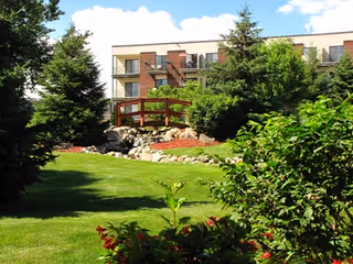 A landscaped outdoor area with green grass, bushes, and trees. A small wooden bridge crosses over a rocky stream or dry creek bed. In the background, there is a multi-story building with balconies under a partly cloudy sky.