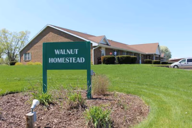 Exterior view of a single-story brick building with a green sign in the foreground that reads 'WALNUT HOMESTEAD'. The building is surrounded by a well-maintained lawn and some bushes, with a clear blue sky above.