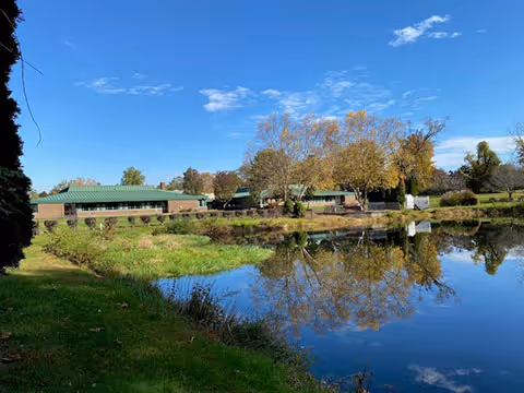 A serene outdoor scene at Masonicare at Bishop Wicke Health Center featuring a calm pond reflecting trees with autumn foliage and a building with a green roof under a clear blue sky.