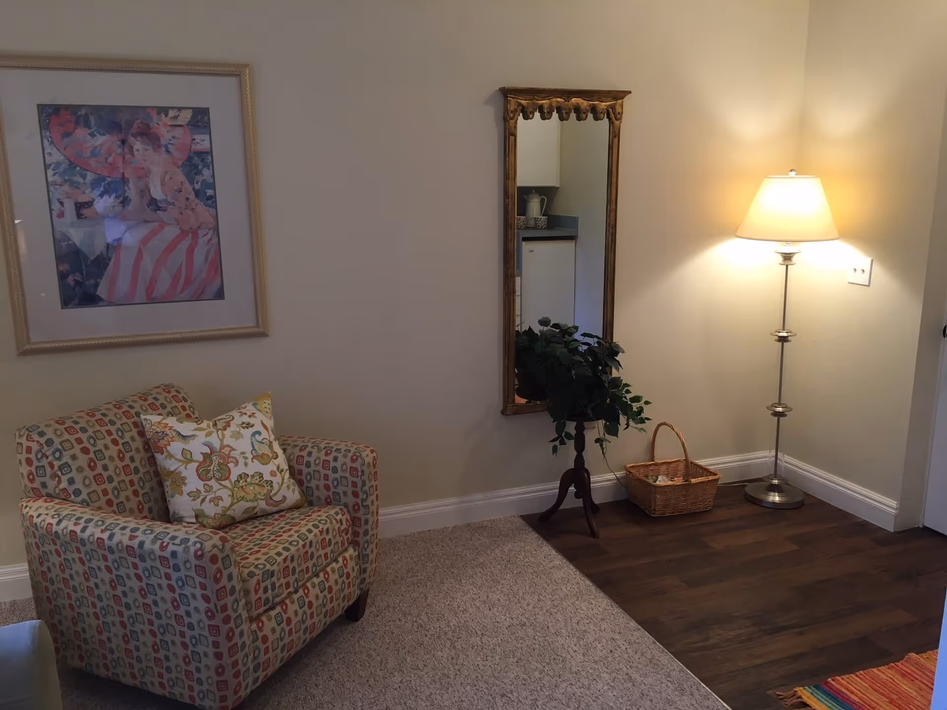 A cozy corner of a living room featuring a patterned armchair with a decorative pillow, a tall floor lamp emitting warm light, a small wooden stand with a potted plant, a wicker basket on the floor, and a framed artwork on the wall. The floor transitions from carpet to wood, and a tall mirror is mounted on the wall.