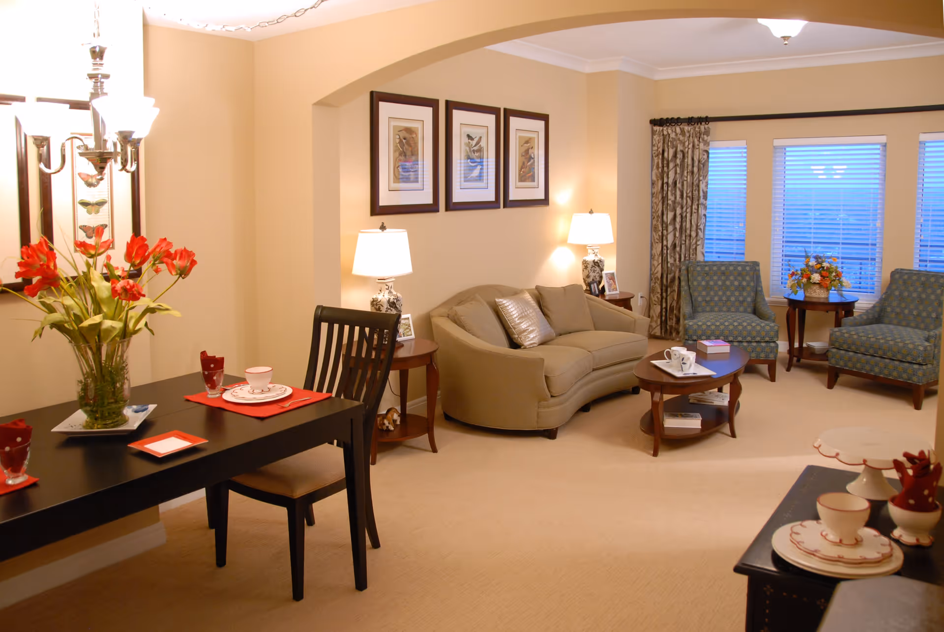 A cozy living room and dining area with beige walls and carpet. The living room features a beige sofa with two decorative pillows, two patterned armchairs, a coffee table with books and a tray, and side tables with lamps and framed photos. The dining area has a black table set with red placemats, white dishes, and a vase of red flowers. Three framed pictures hang on the wall above the sofa, and windows with blinds and patterned curtains let in natural light.