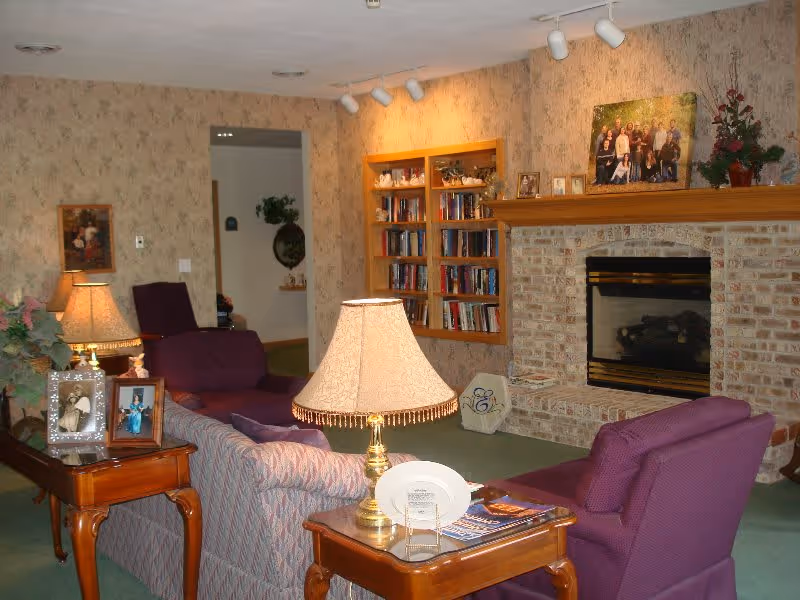 Cozy living room in Eastland Retirement Community featuring purple upholstered chairs and sofa, a wooden side table with a lamp and framed photos, a brick fireplace with a wooden mantle displaying family photos and a plant, and a built-in bookshelf filled with books.