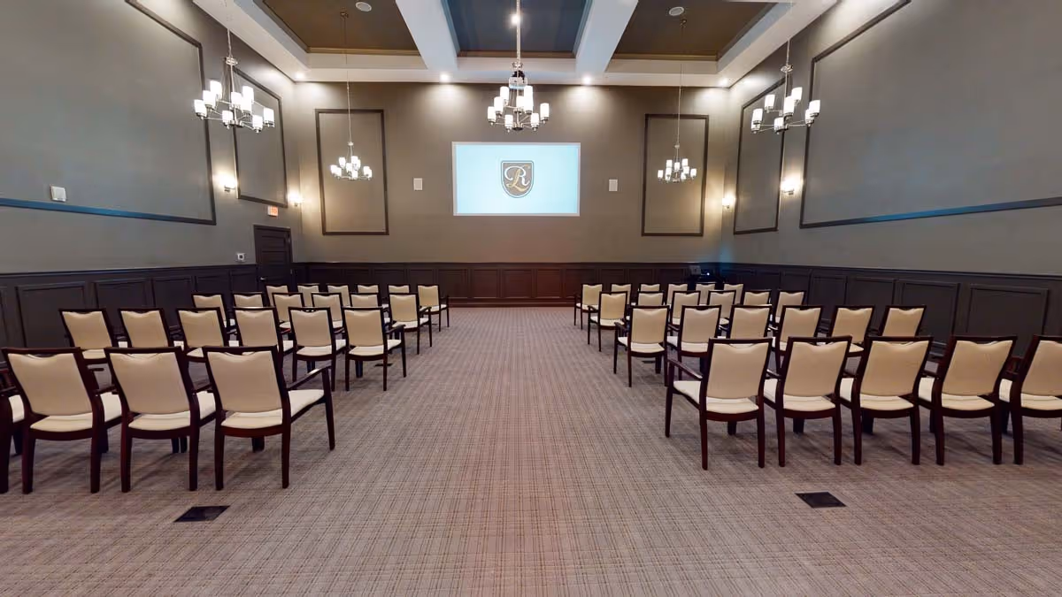 A large conference or meeting room with rows of beige cushioned chairs arranged facing a wall with a projector screen displaying a logo. The room has high ceilings with multiple chandeliers and wall sconces providing lighting. The walls are painted gray with dark wood paneling on the lower half.