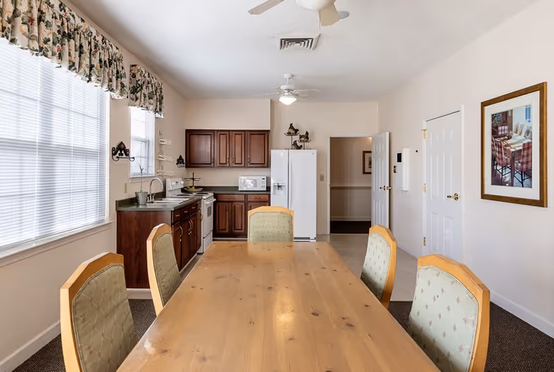 A bright kitchen and dining area in an assisted living facility featuring a wooden dining table with six cushioned chairs. The kitchen has dark wooden cabinets, a white refrigerator, microwave, stove, and a sink under two windows with floral valances. The walls are light-colored with a framed picture, and there are ceiling fans with lights.