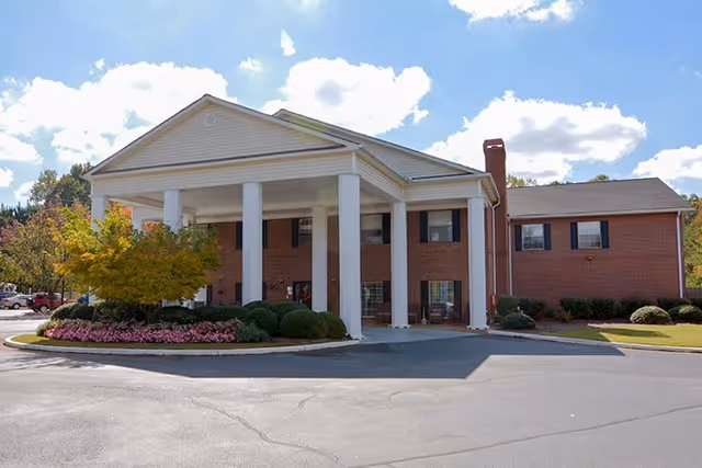 Front entrance of a brick senior living building with large white columns, a covered porte-cochère, landscaping, and a paved driveway.