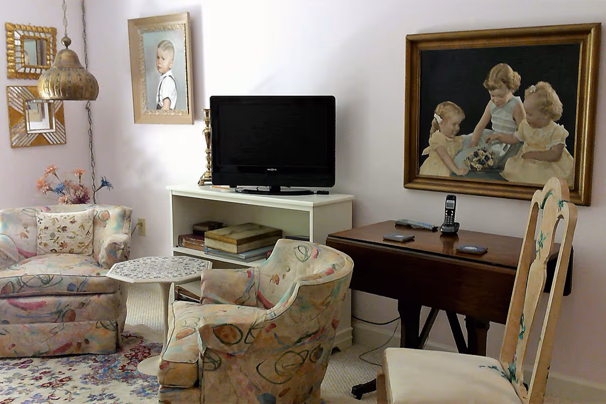 A cozy living room with pastel floral upholstered armchairs and a small white table. A TV sits on a white shelf with books underneath. On the wall are framed paintings, including one of three young girls and another of a young boy. A wooden table with a cordless phone and remote controls is next to the chairs.