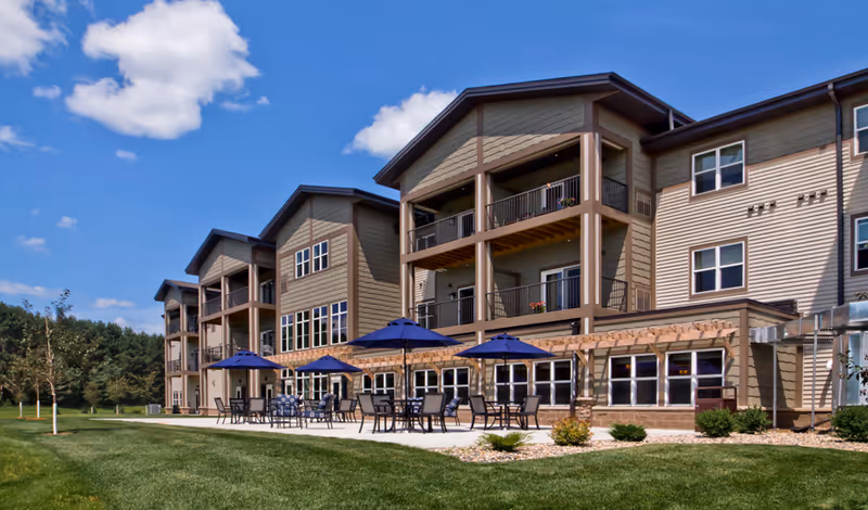 Exterior view of a multi-story senior living facility with beige siding and multiple balconies. In front of the building, there is a patio area with tables, chairs, and blue umbrellas. The area is surrounded by green grass and small landscaped bushes under a partly cloudy blue sky.