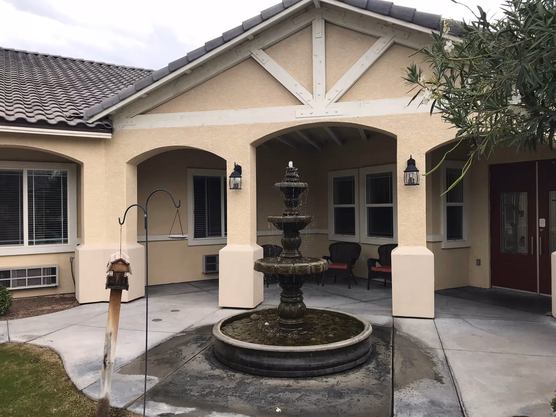 Outdoor courtyard area of Visions Assisted Living featuring a three-tiered water fountain in the center, surrounded by a concrete walkway. The building has beige stucco walls with white trim and a tiled roof. There are two black chairs with red cushions under a covered patio area, windows with blinds, and wall-mounted lantern-style lights. A bird feeder hangs on a metal hook near the fountain, and some greenery is visible on the right side.