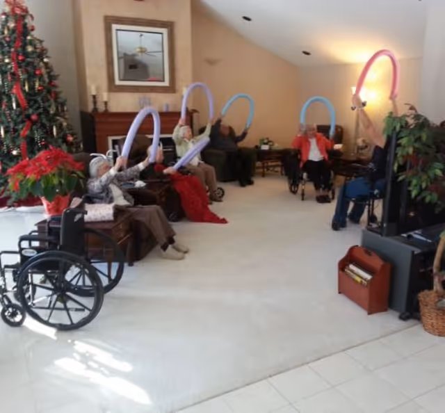 A group of elderly individuals seated in a living room area, some in wheelchairs, holding colorful exercise bands above their heads. The room features a decorated Christmas tree, a fireplace with a mirror above it, and a television on a stand. The atmosphere appears warm and engaging.
