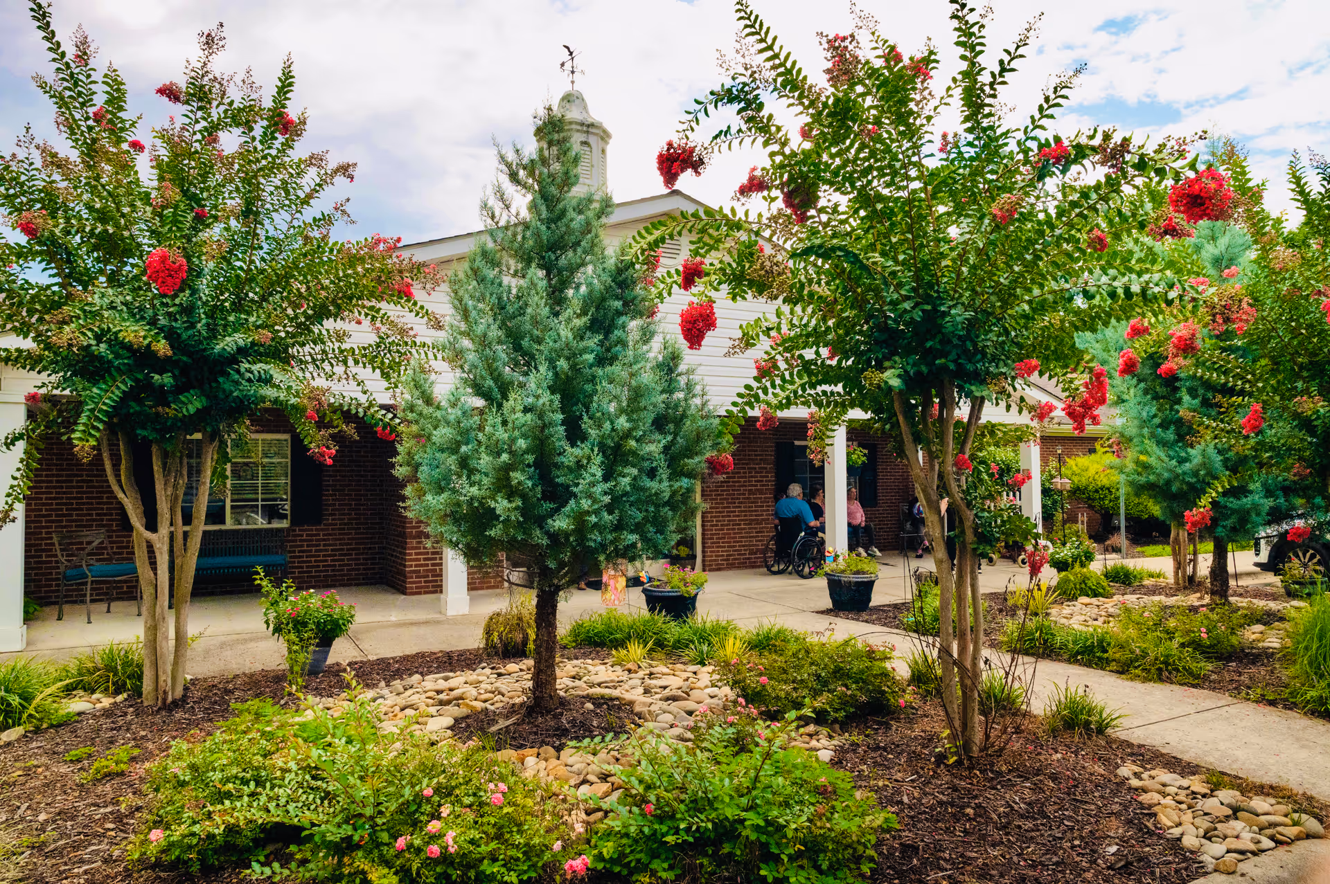 Outdoor view of Cambridge House facility showing a garden with small trees and flowering plants in front of a brick building with a covered porch. Two people are sitting on the porch, one in a wheelchair. The sky is partly cloudy.