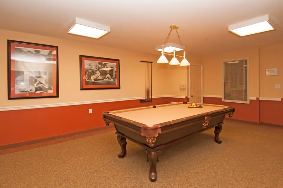 Interior room with a pool table in the center, beige walls with a red lower half, two framed black and white pictures on the wall, a window with blinds, and a ceiling light fixture above the pool table.