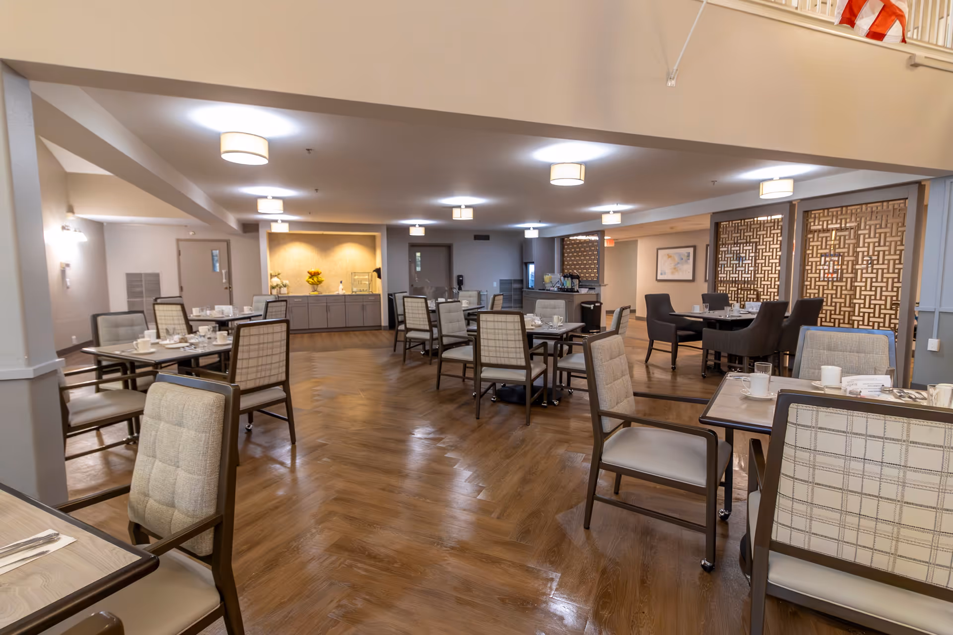 A spacious dining room with multiple tables and chairs arranged neatly. The room features wooden flooring, modern ceiling lights, and a decorative partition on one side. Tables are set with cups, glasses, and napkins, ready for dining. The overall ambiance is warm and inviting.