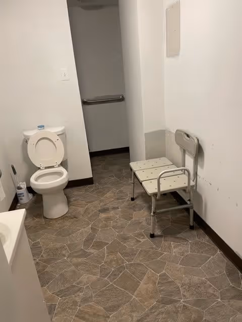 A bathroom with a white toilet with the lid open, a small white sink partially visible on the left, a white shower chair with armrests on the right, and a grab bar on the wall in the background. The floor has a stone-patterned tile design and the walls are plain white.
