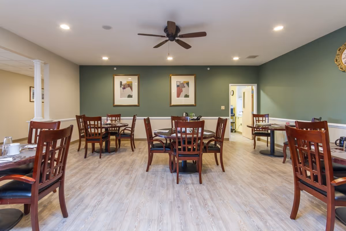 Dining room with several wooden tables and chairs arranged on a light wood floor. The walls are painted green and beige with two framed abstract paintings and a clock. A ceiling fan and recessed lights are visible on the ceiling. An open doorway leads to another room.