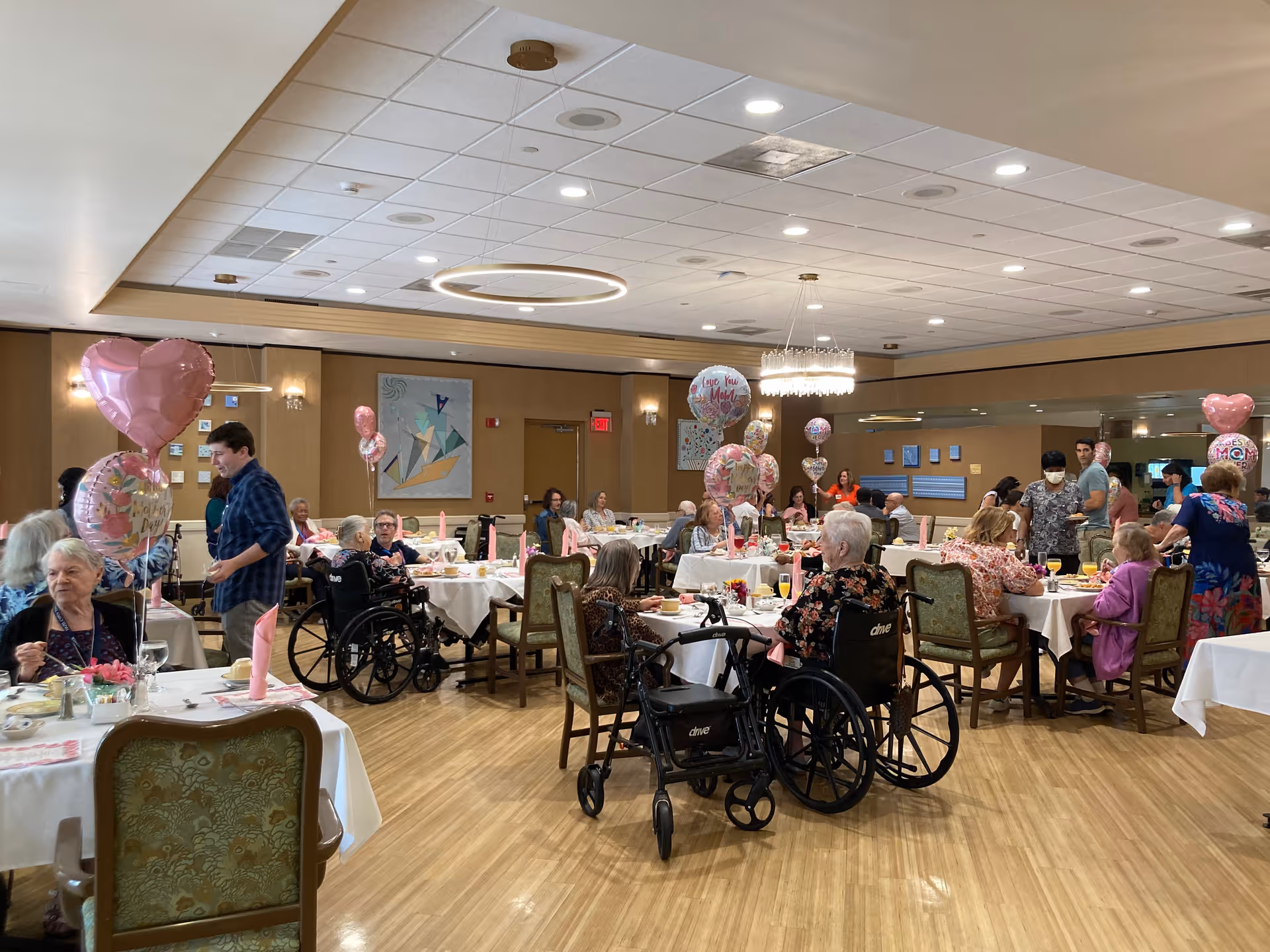 A large dining room filled with elderly residents seated at tables covered with white tablecloths, decorated with pink heart-shaped balloons and floral centerpieces. Some residents are in wheelchairs, and caregivers or visitors are standing and interacting with them. The room has wooden floors, beige walls, and modern ceiling lights.