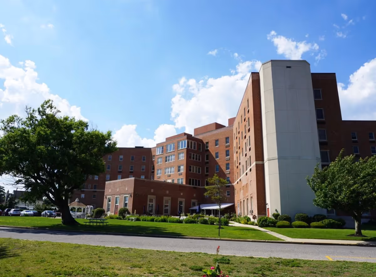 Exterior view of a multi-story brick building with several windows, surrounded by green lawns, trees, and a clear blue sky with some clouds.