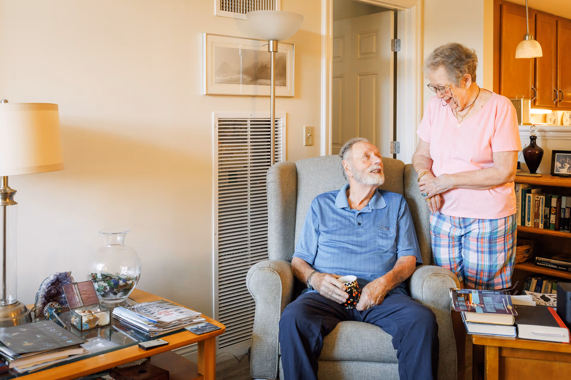 An elderly man sitting in a comfortable armchair holding a coffee mug, looking up and smiling at an elderly woman standing beside him in a cozy living room with bookshelves, lamps, and a side table with magazines and decorative items.